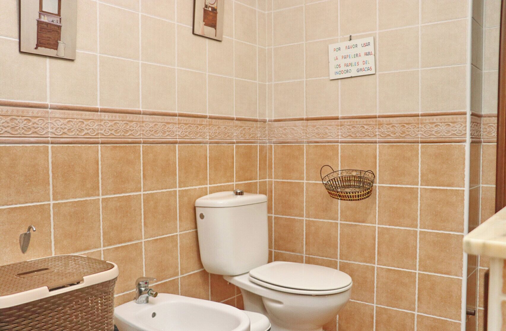 Een badkamer met toilet, bidet en wastafel. Er is een decoratieve tegelrand en een bordje aan de muur.