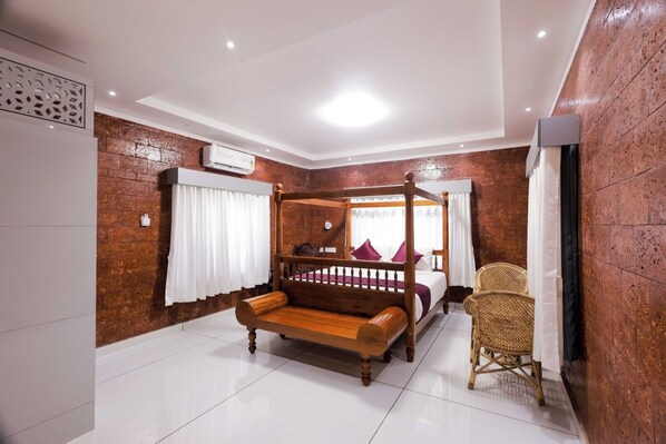 A bedroom with a four-poster bed, a wooden bench, a wicker chair, and a brick wall.