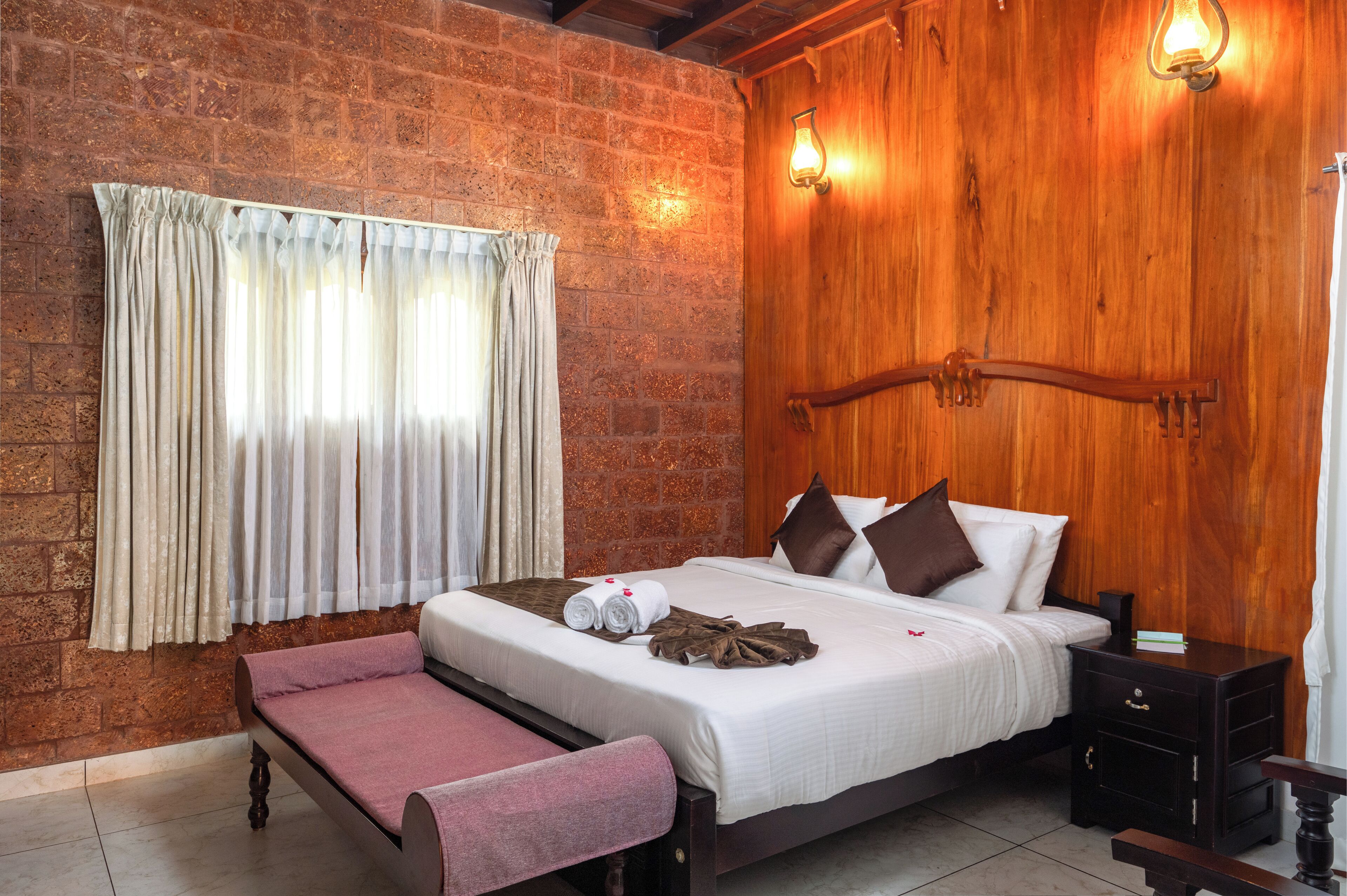A hotel room with a brick wall, wooden headboard, a bed with white linens, a bench, and a nightstand.