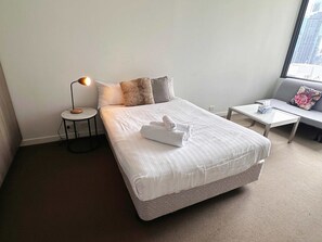 A neatly made bed with white linens and a folded towel, a small round table with a lamp, a sofa with a floral pillow, and a window with a city view. - Cozy Studio Apt Near Southern Cross & City Views (Melbourne)