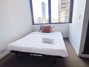Una cama individual con ropa blanca y una toalla enrollada, una ventana con vista a edificios altos y una pared blanca lisa.