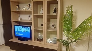 Una sala de estar con televisor en un mueble bajo, un aparador de madera con puertas de vidrio, una planta en maceta y un sofá con almohadones decorativos.