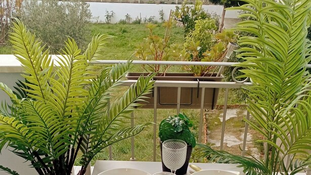 Mesa de comedor servida con platos, vasos y cubiertos en un balcón con plantas en macetas y vistas del entorno.