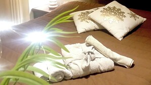 Una cama bien hecha con una colcha marrón, dos almohadas decorativas y una toalla blanca enrollada sobre un piso de madera.