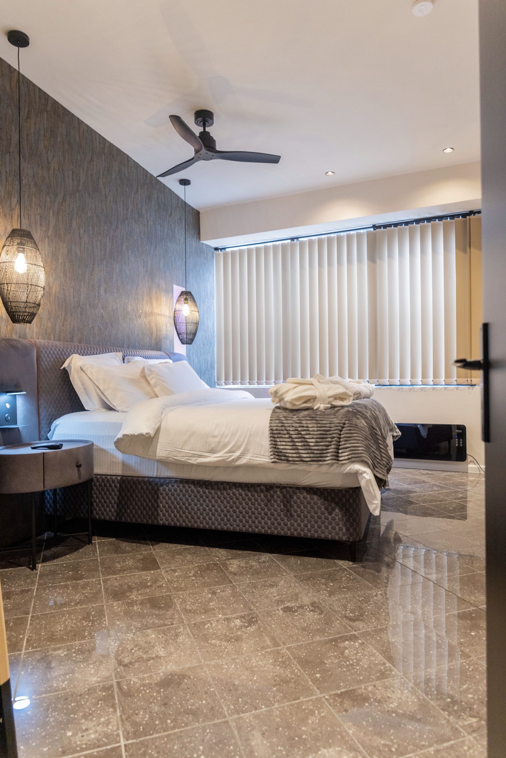 A modern bedroom with a bed, bedside table, lamp, ceiling fan, and a window with blinds.