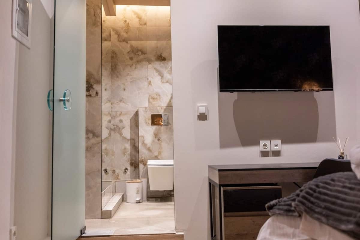Une chambre moderne avec un plancher en bois, un lit, un téléviseur fixé au mur et une salle de bain visible à travers une porte ouverte.