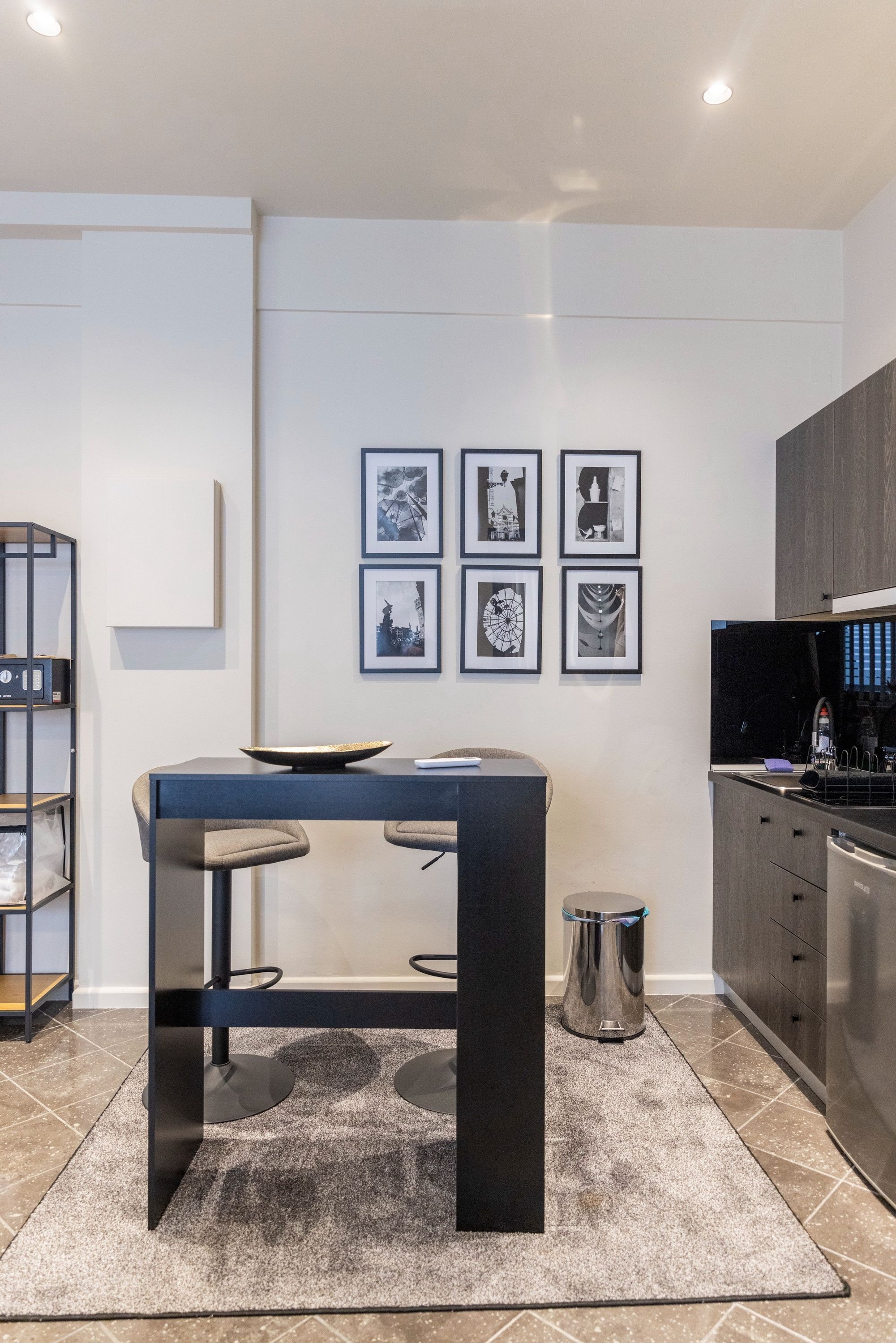 A modern kitchen with a dark countertop, a small table with stools, and a coffee machine. There are framed pictures on the wall and a trash can next to the table.