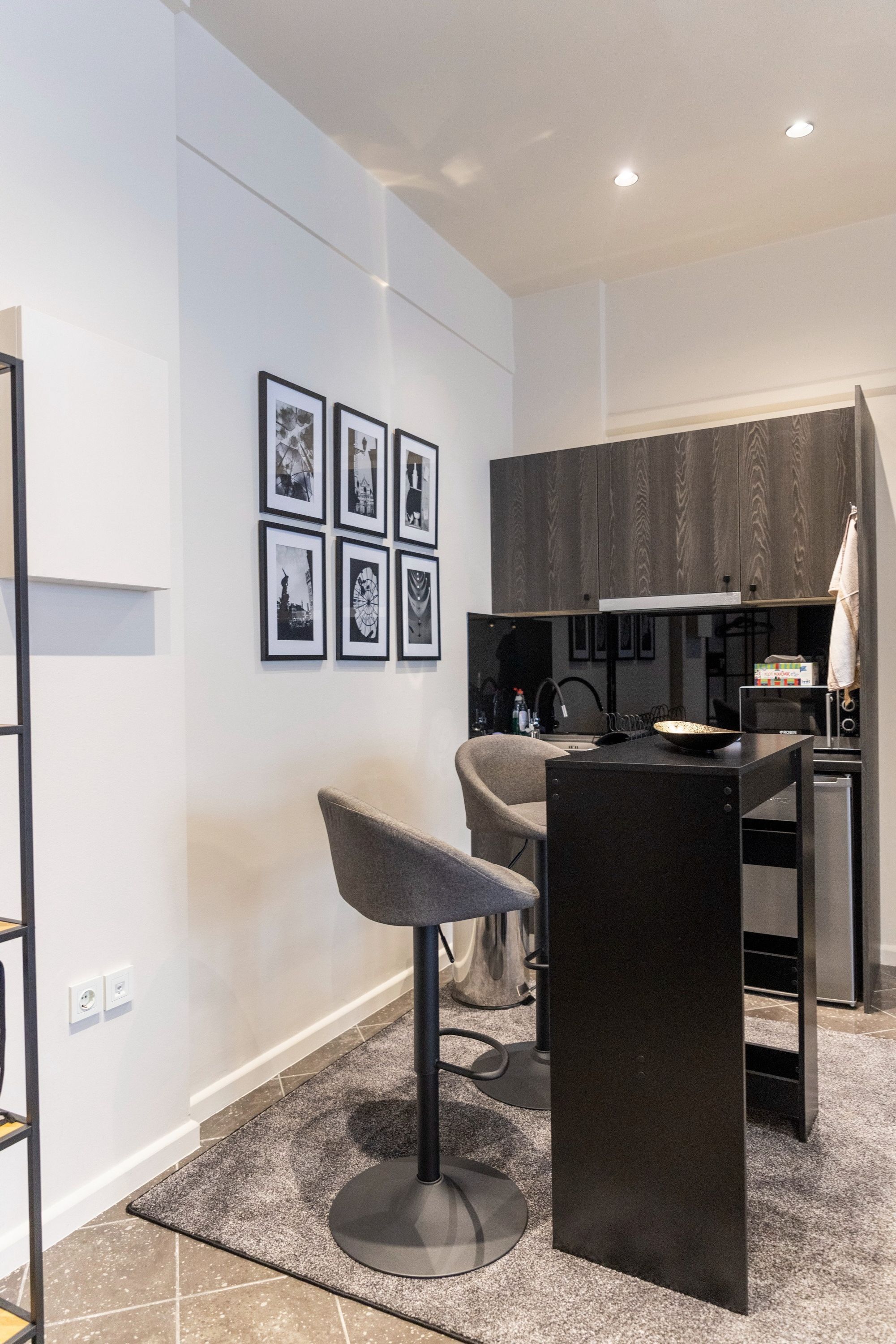 A modern kitchen with a dining area, featuring a black dining table, a grey chair, and a black bar stool. There are framed pictures on the wall and a kitchen island with a sink and countertop.