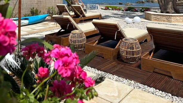 Un espace de détente aménagé sur la plage, avec des chaises longues en bois, un parasol blanc et un pin.