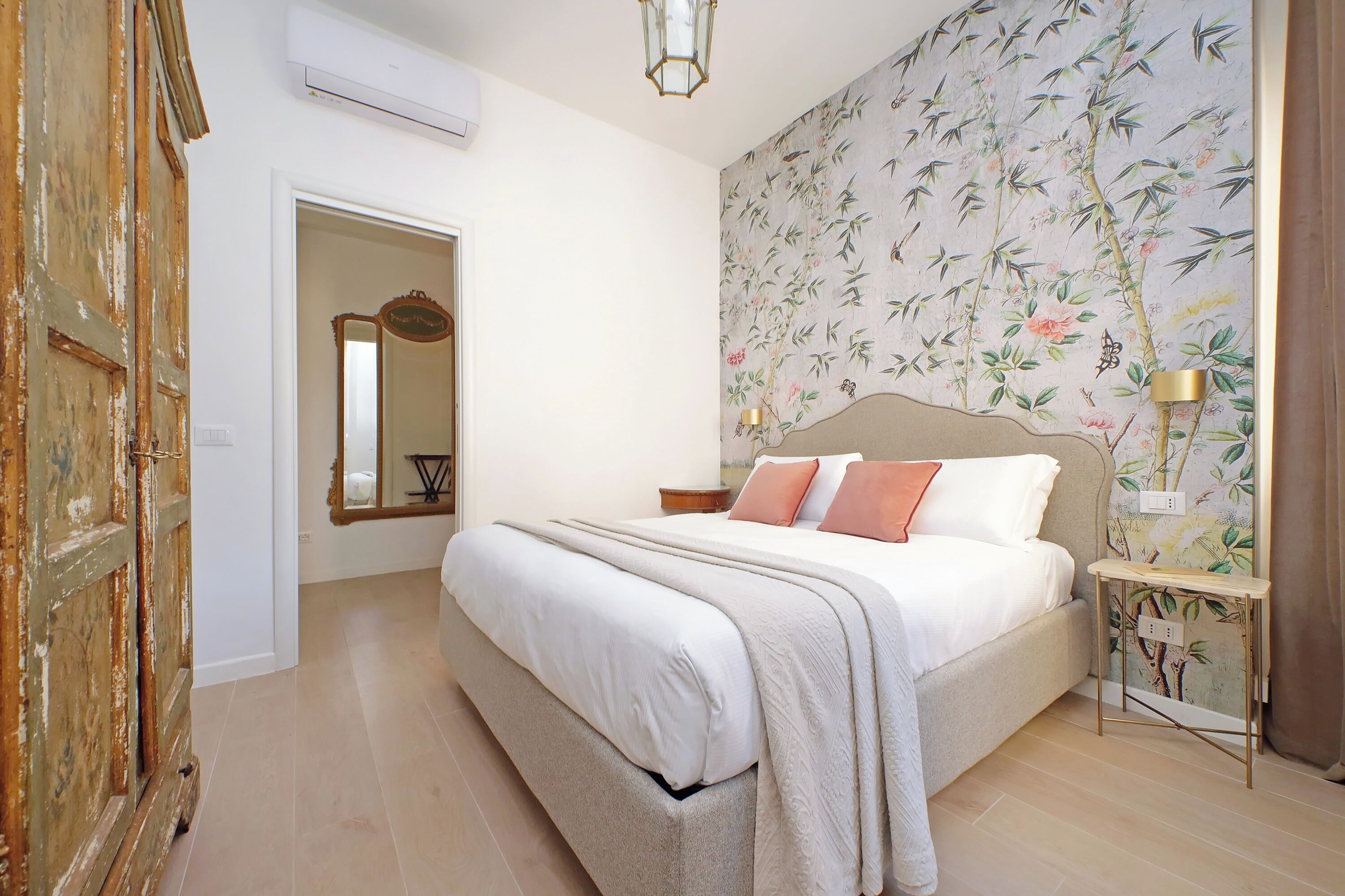 Une chambre à coucher avec un lit, une table de chevet, un miroir et un mur recouvert d’un papier peint à motifs floraux.