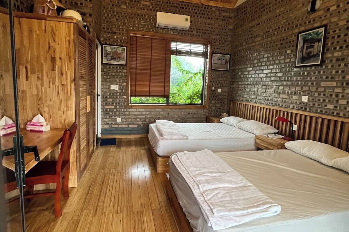 Habitació amb sostre de fusta, ventilador de sostre, finestra amb vista a vegetació, dos llits, escriptori de fusta amb cadires i paret amb dibuix de rajoles.