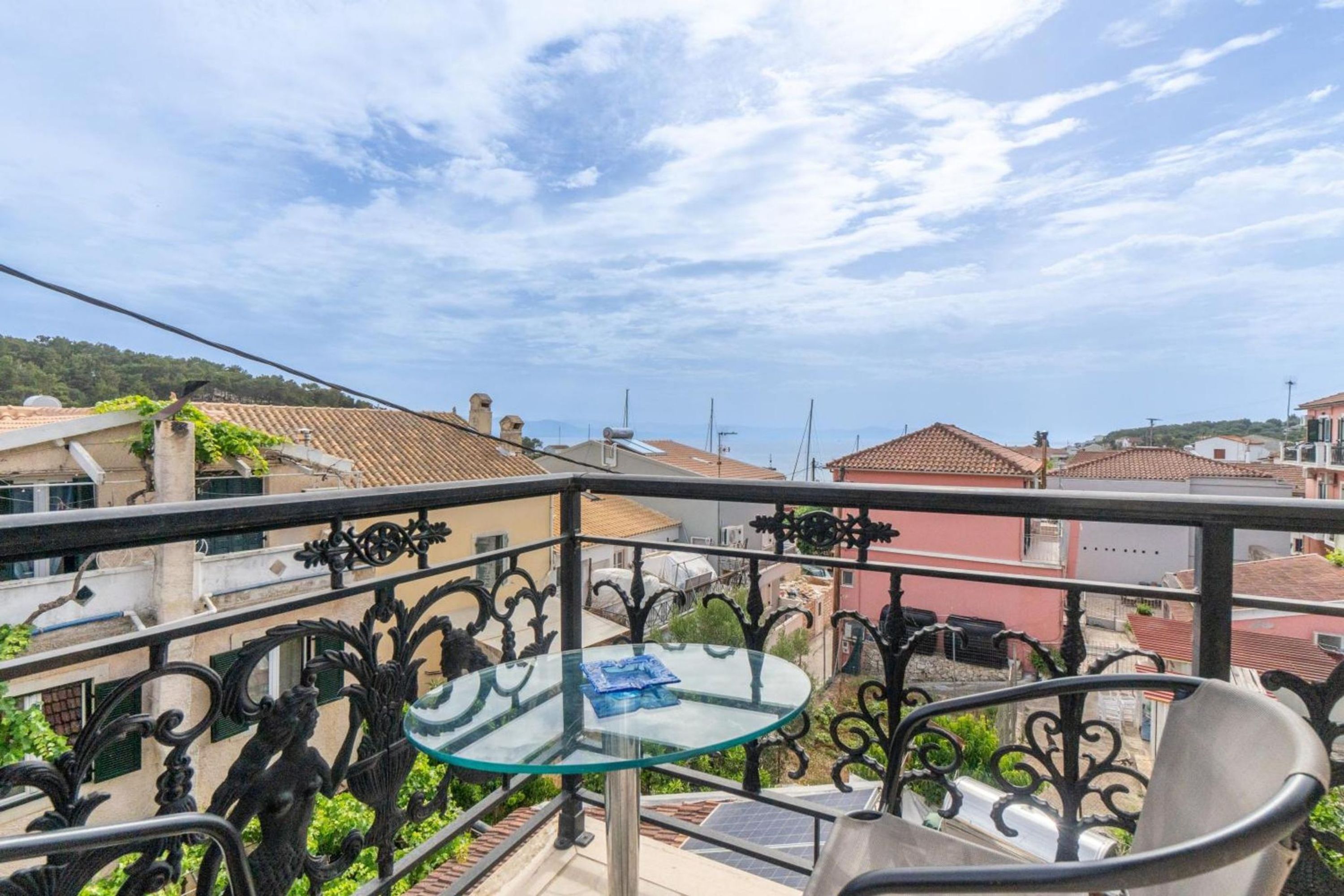Varanda com mesa de vidro e grades de metal, com vista para uma área residencial com telhados de telha vermelha.