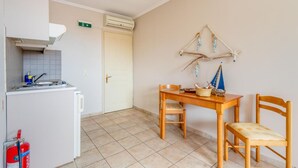 A compact kitchen with a white refrigerator, a stove, and a sink. There is a fire extinguisher on the floor. A wooden table with chairs is positioned against the wall, and a small decorative sailboat hangs above it. - Paxi Apt in Gaios With 1 Bedrooms and 1 Bathrooms (Paxos)