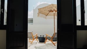 Puerta abierta que da a un balcón con un paraguas de mimbre, dos sillas de madera y una mesa redonda.