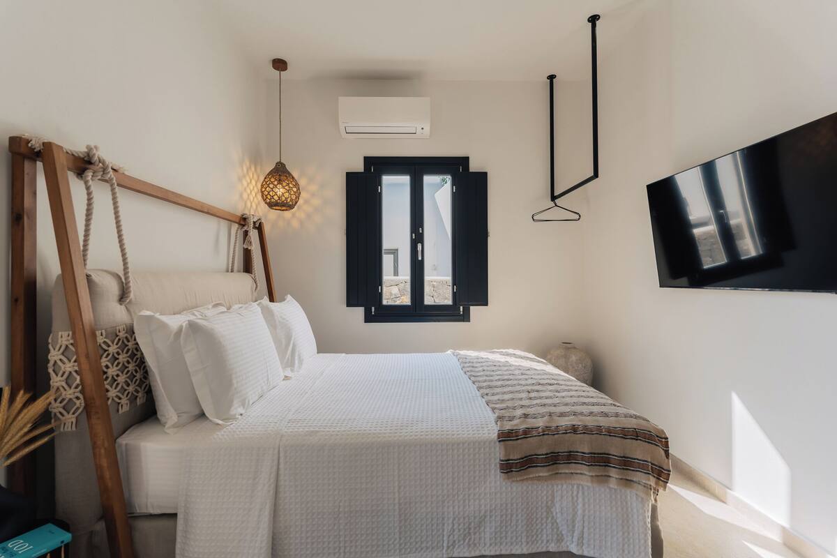 A bedroom with a bed, a hanging light fixture, a TV mounted on the wall, and a window with shutters.