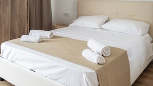 Un letto rifatto con lenzuola bianche e copriletto color beige, un comodino in legno con un piccolo tavolo, e una parete liscia con un interruttore.