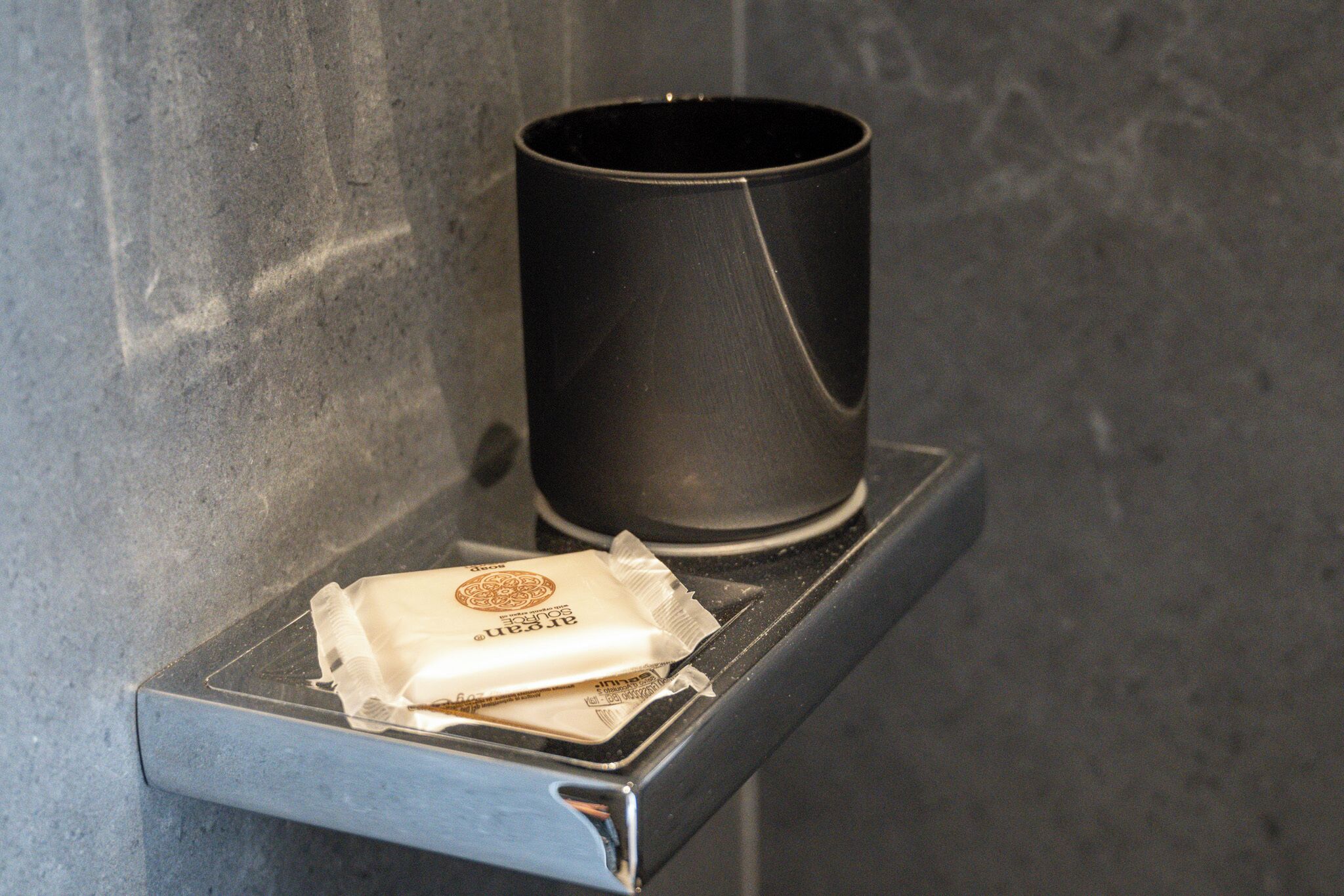 Un objeto cilíndrico negro sobre una superficie metálica, posiblemente un recipiente o una taza, con un paquete blanco parcialmente visible debajo.
