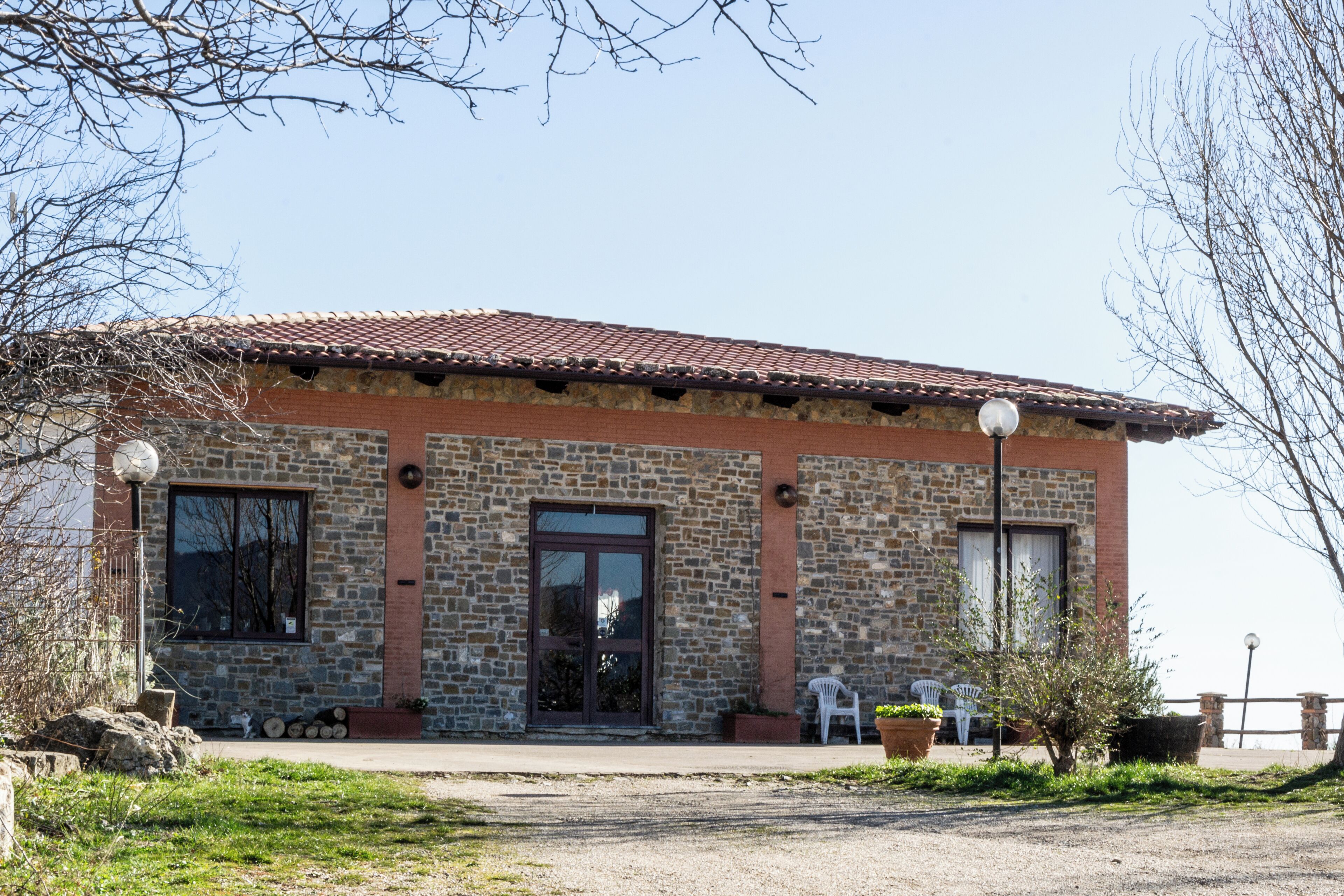 A stone building with a tiled roof, a front door, and windows. There are outdoor seating areas with chairs and potted plants.
