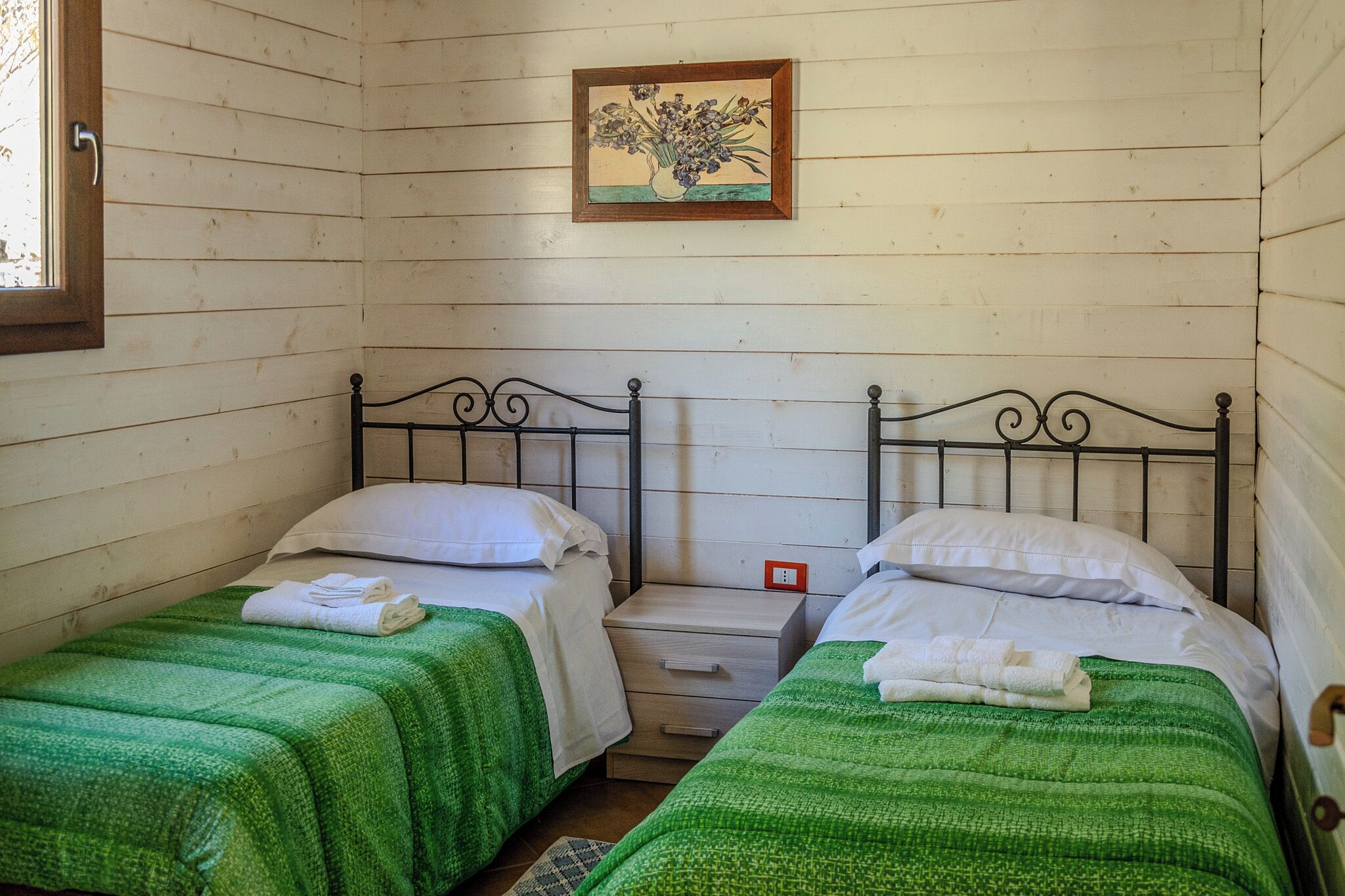 Une petite chambre agréable, dotée de deux lits simples, chacun recouvert d’une couverture verte et agrémenté d’oreillers blancs. Une petite table de chevet en bois est placée entre les lits, et un tableau représentant des fleurs est accroché au mur, au-dessus des lits.