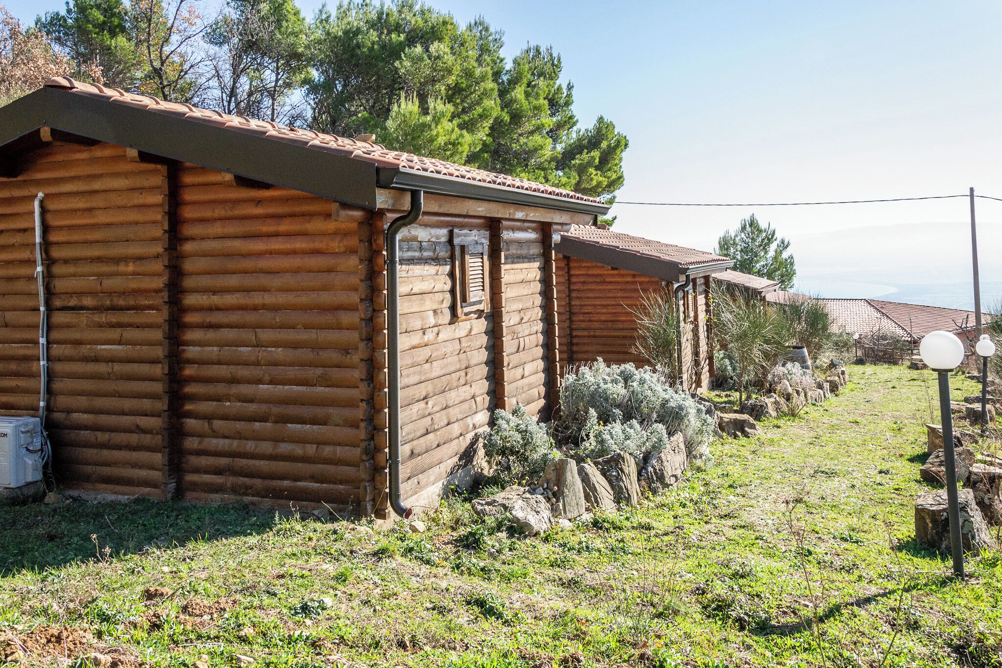Una cabaña rústica de madera con techo de tejas, rodeada de césped, rocas y árboles.