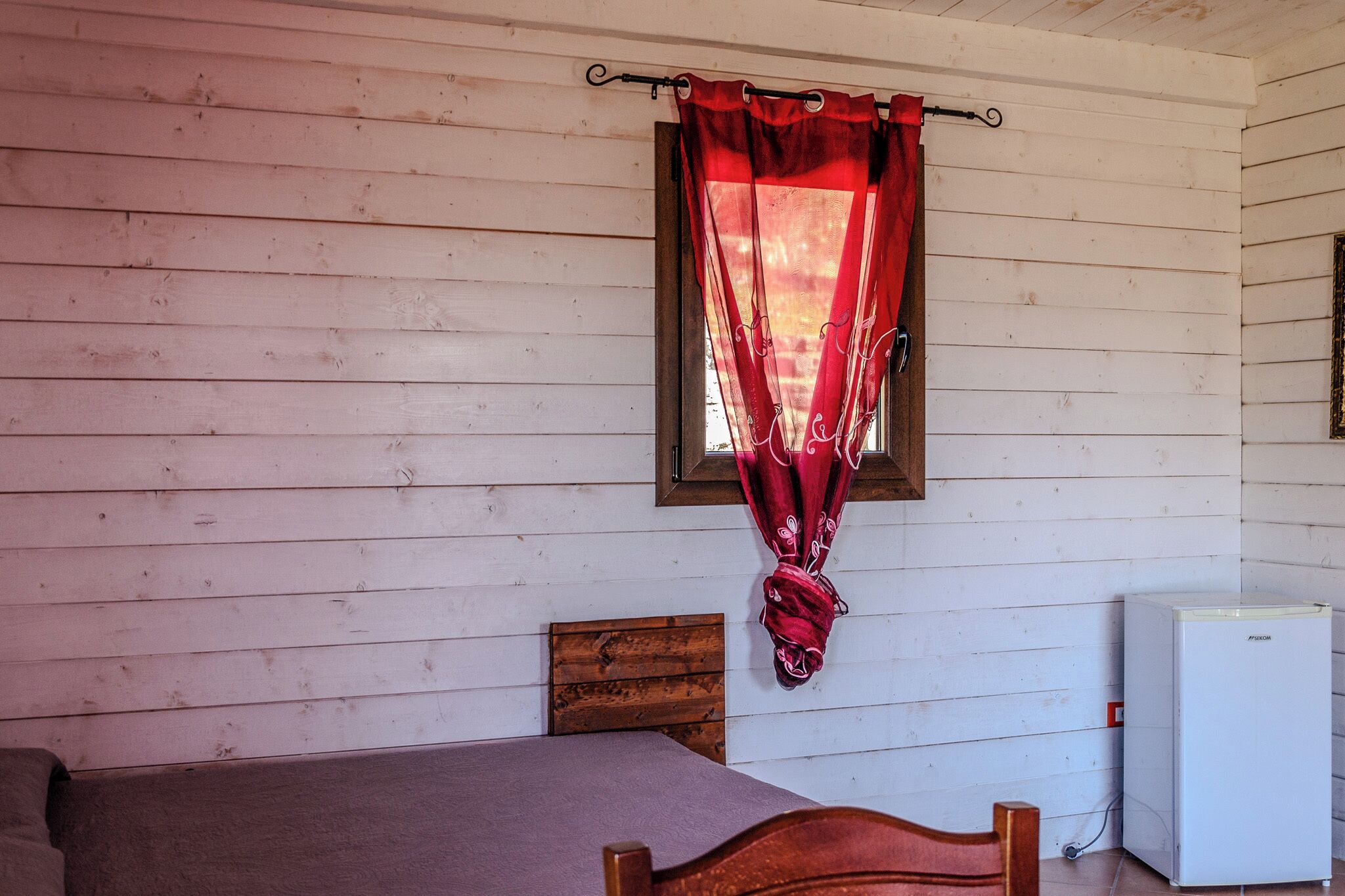 A room with a bed, a wooden headboard, a window with a red curtain, and a white refrigerator.