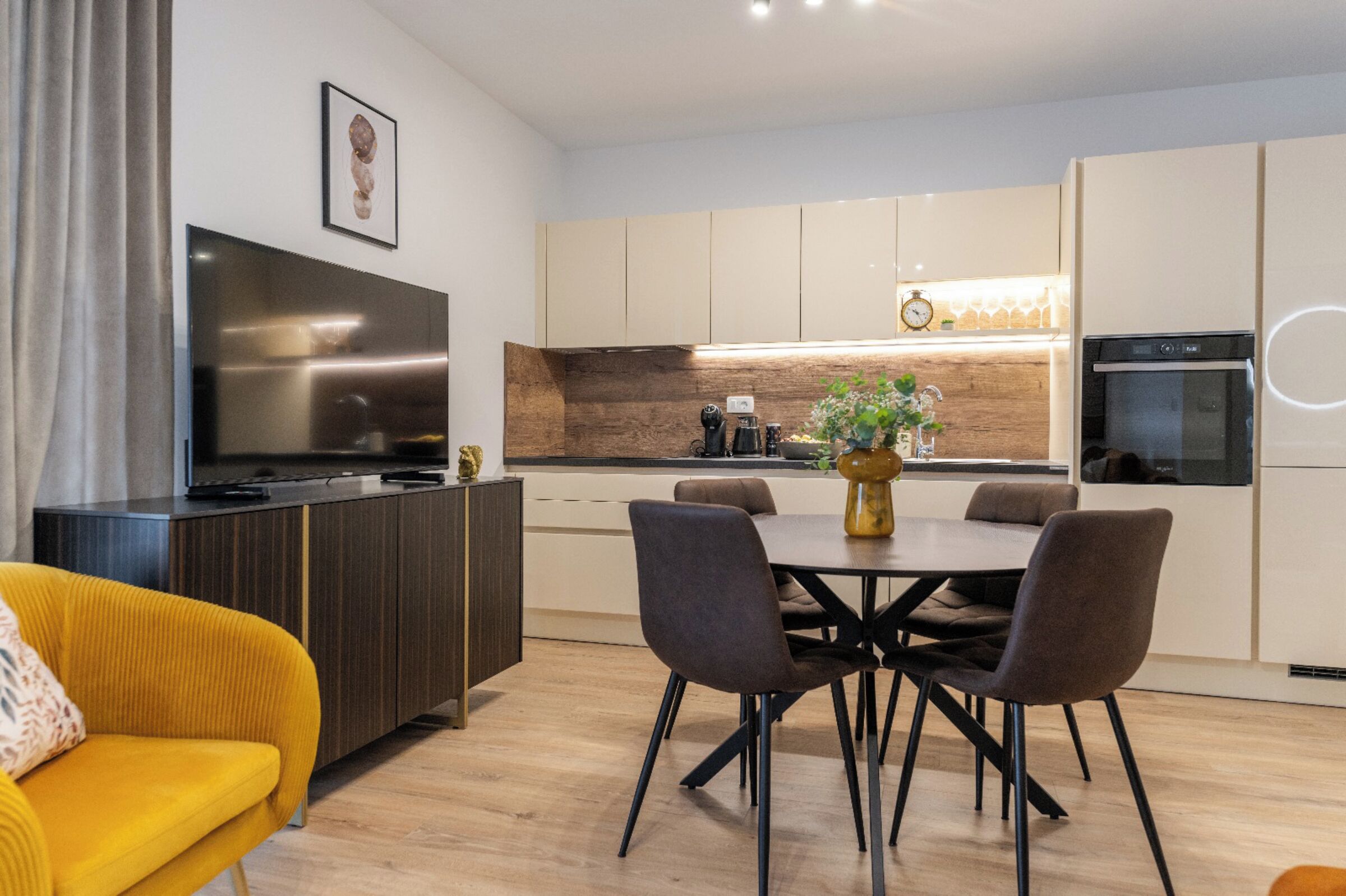 A modern kitchen with a dining area, a flat-screen TV, and a yellow sofa.
