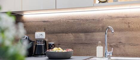 Een moderne keuken met een houten spatwand, een koffiezetapparaat, een fruitschaal, een gootsteen en een klok aan de muur.