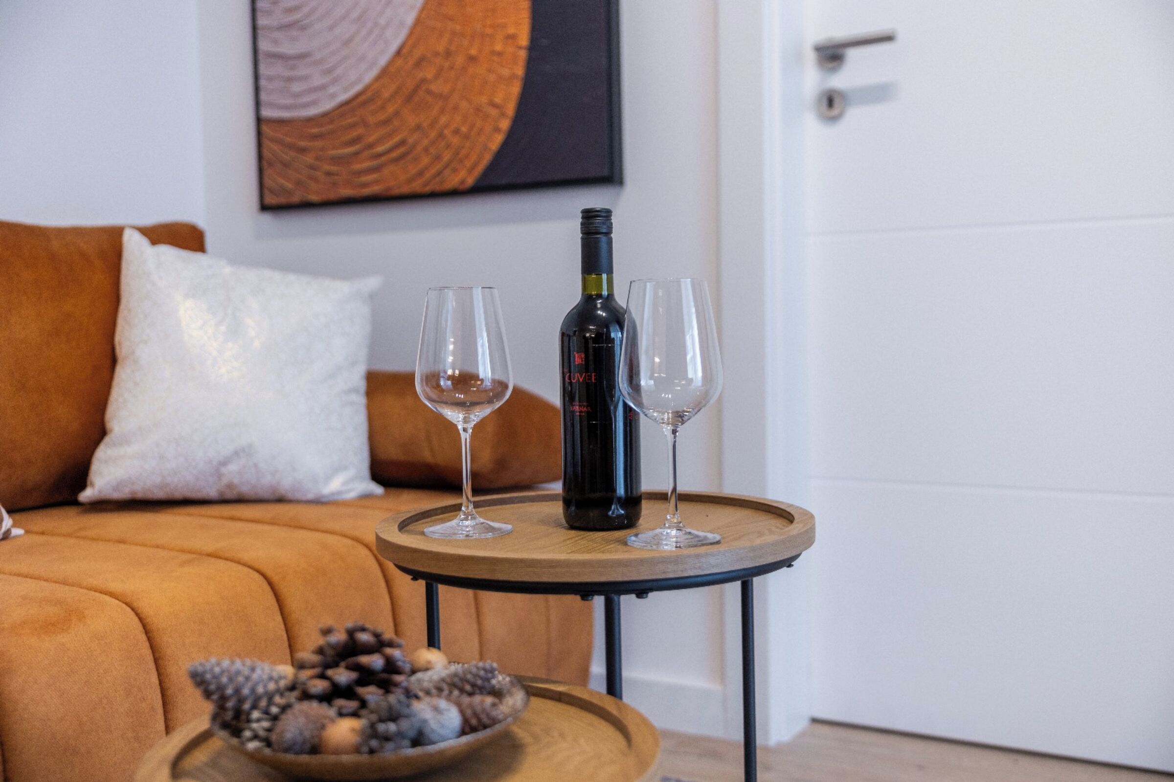 Une bouteille de vin sur un plateau, accompagnée de deux verres, d’un bol de noix, et d’un canapé marron avec un coussin blanc, dans une pièce avec une porte blanche et un tableau au mur.