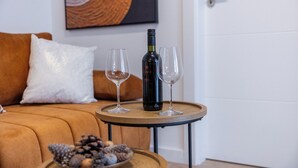 Une bouteille de vin sur un plateau, accompagnée de deux verres, d’un bol de noix, et d’un canapé marron avec un coussin blanc, dans une pièce avec une porte blanche et un tableau au mur.