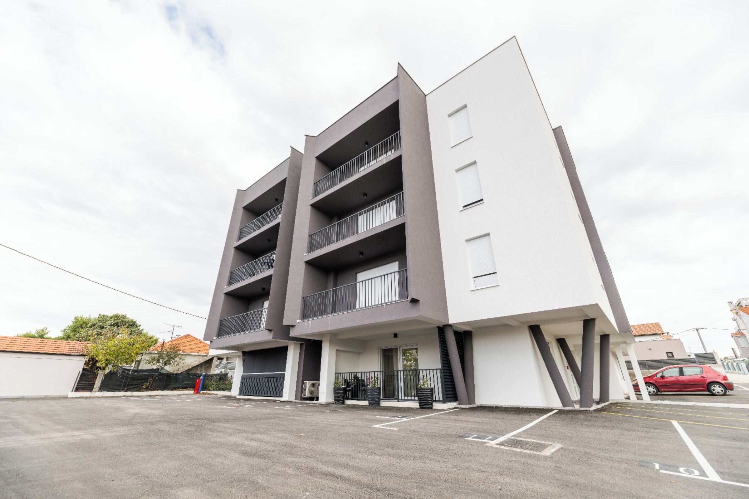 Un edificio moderno de varios pisos con balcones, un estacionamiento y un coche rojo aparcado cerca.