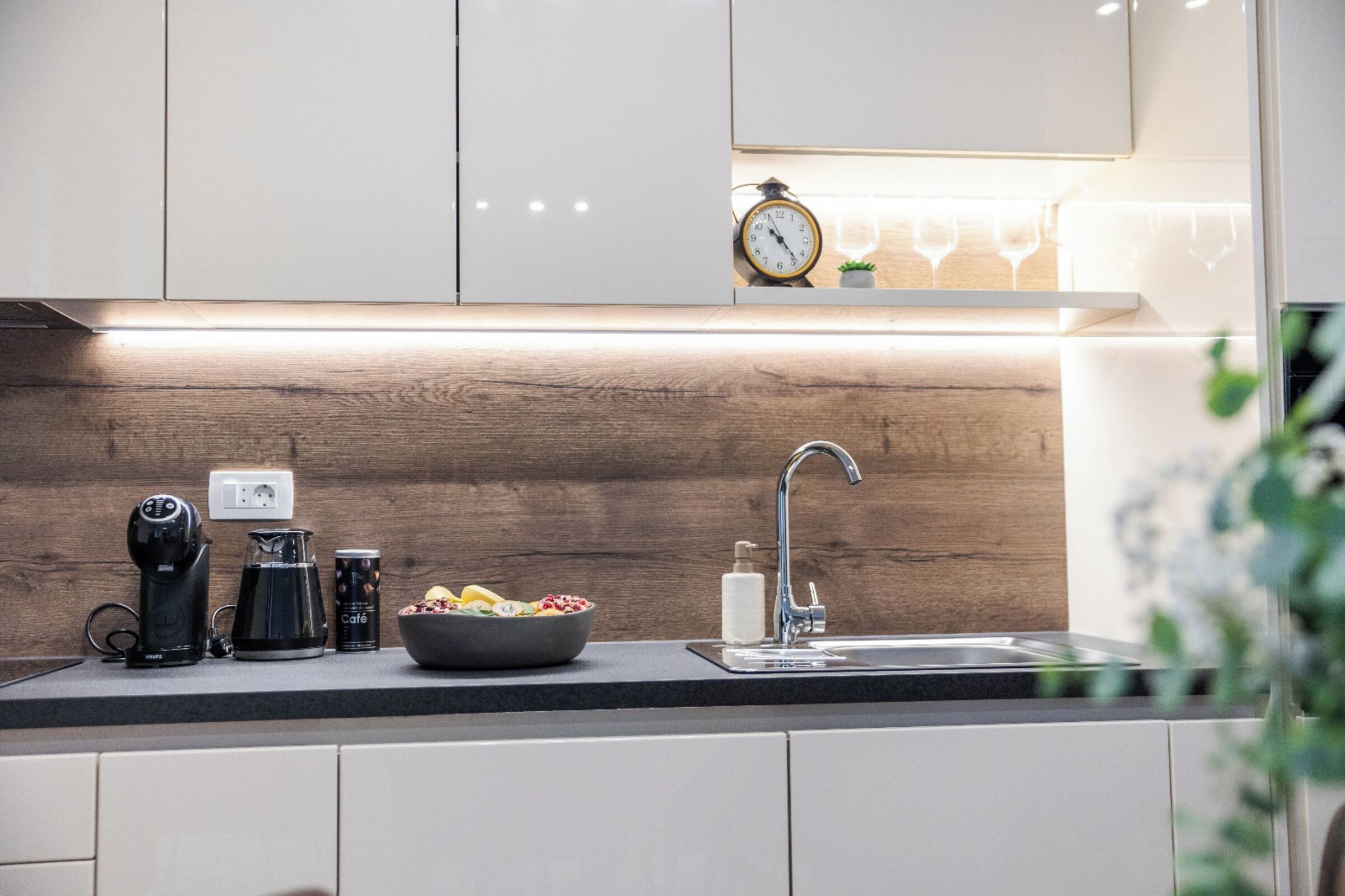 A modern kitchen with white cabinets, a wooden backsplash, a coffee maker, a kettle, a bowl of fruit, a sink, and a clock on the wall.