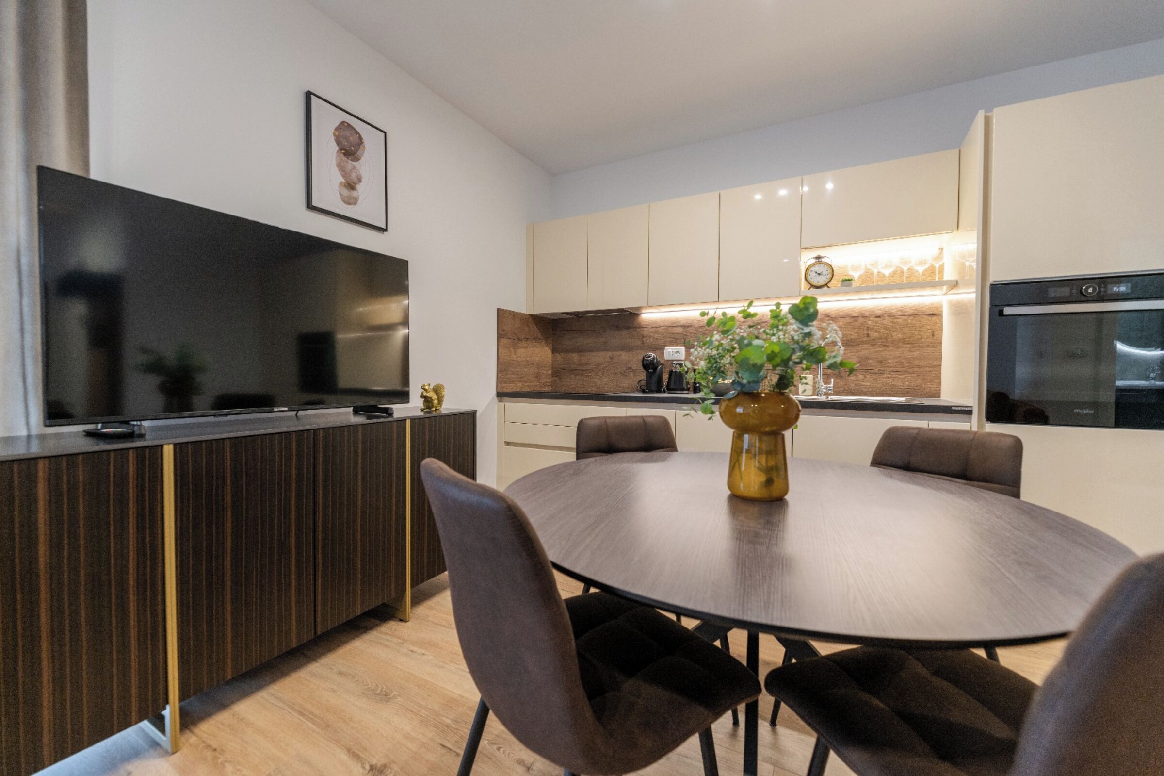 A modern kitchen with a dining area, featuring a round table, chairs, a vase with flowers, and a clock on the wall.