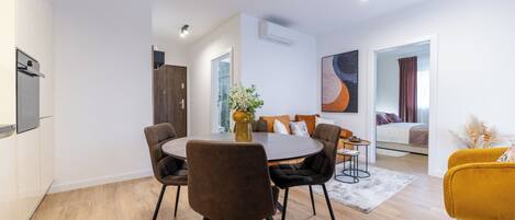 A modern living room with a round dining table, comfortable seating, a sofa, a bed, and a large window.