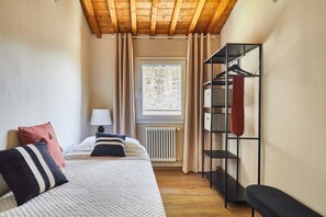 Un dormitorio con cama, mesita de noche, lámpara, radiador, ventana con cortinas y perchero.