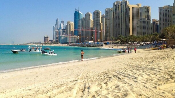 A beach with a city skyline in the background, including high-rise buildings and a marina with boats. - Alharaki Capsule  (Dubai)