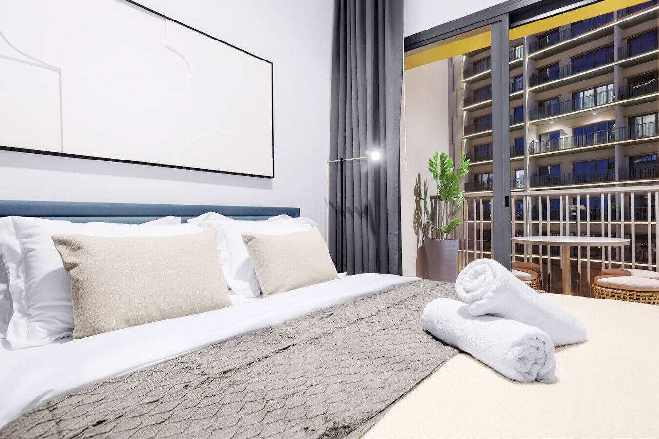 Ein modernes Hotelzimmer mit einem großen Bett, einem grauen Teppich mit Struktur, einem ordentlich gefalteten Handtuch und Blick auf die Außenansicht des Gebäudes durch ein großes Fenster.