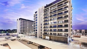 Et moderne, flereetagers hotel med en flad facade, altaner på hver etage og en parkeringsplads foran.
