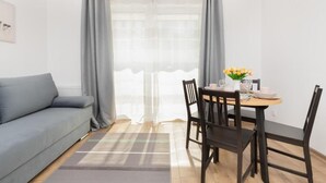 A modern living room with a grey sofa, a wooden dining table with chairs, a small potted plant, and a window with grey curtains.