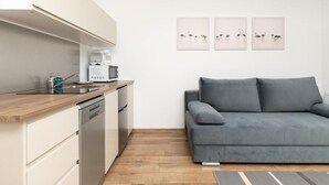 A modern kitchen with a built-in oven, a coffee maker, and a dishwasher. There is a grey sofa against the wall and three framed pictures above it.