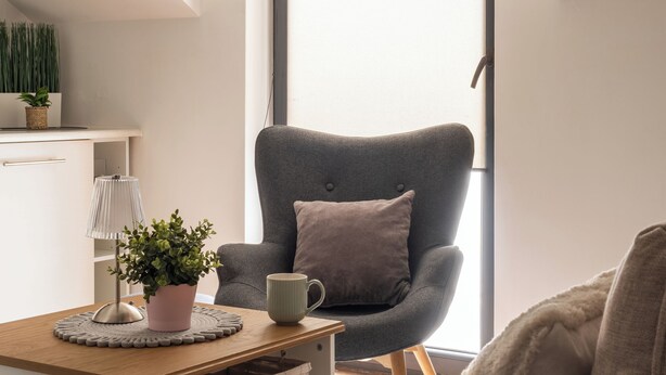 Ein modernes, minimalistisches Zimmer mit einem grauen Sessel, einem kleinen Tisch mit Lampe und Pflanze sowie einem großen Fenster für natürliches Licht.