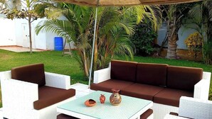 Ein möblierter Patio im Freien mit weißen Korbmöbeln, einem Glastisch und einem großen Sonnenschirm.