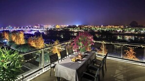 Ένας χώρος τραπεζαρίας με ένα τραπέζι στρωμένο για δύο άτομα, με θέα σε ένα ποτάμι και τα φώτα της πόλης στο βάθος.