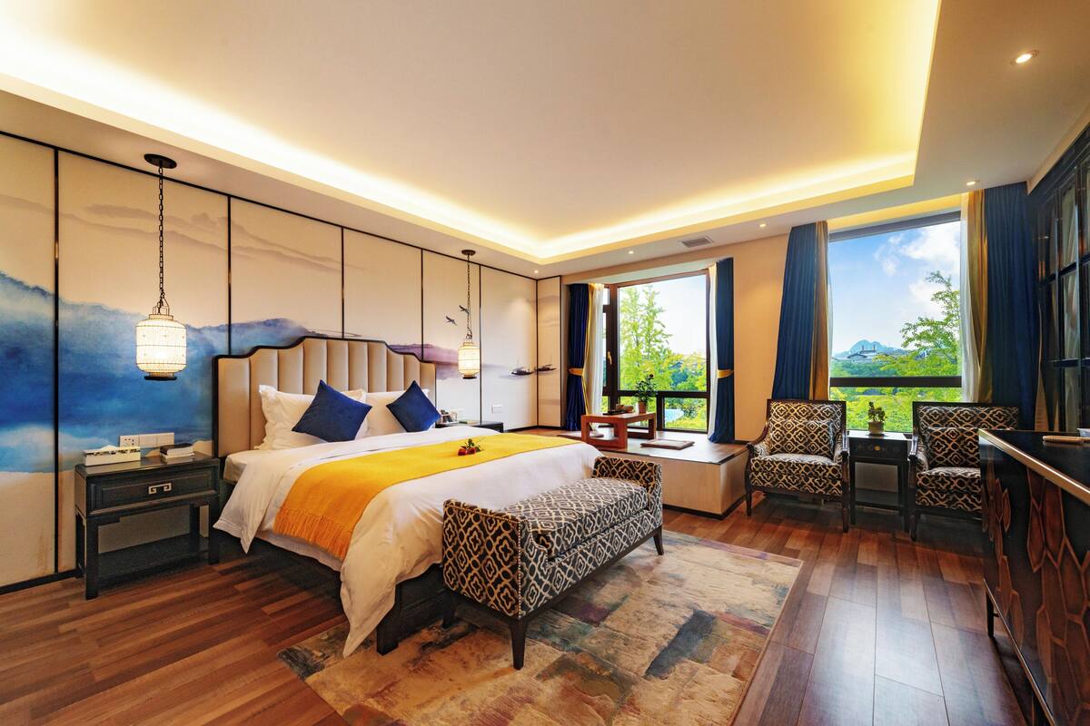Una habitación de hotel moderna con una cama grande, un escritorio, dos sillones estampados y vistas al exterior.