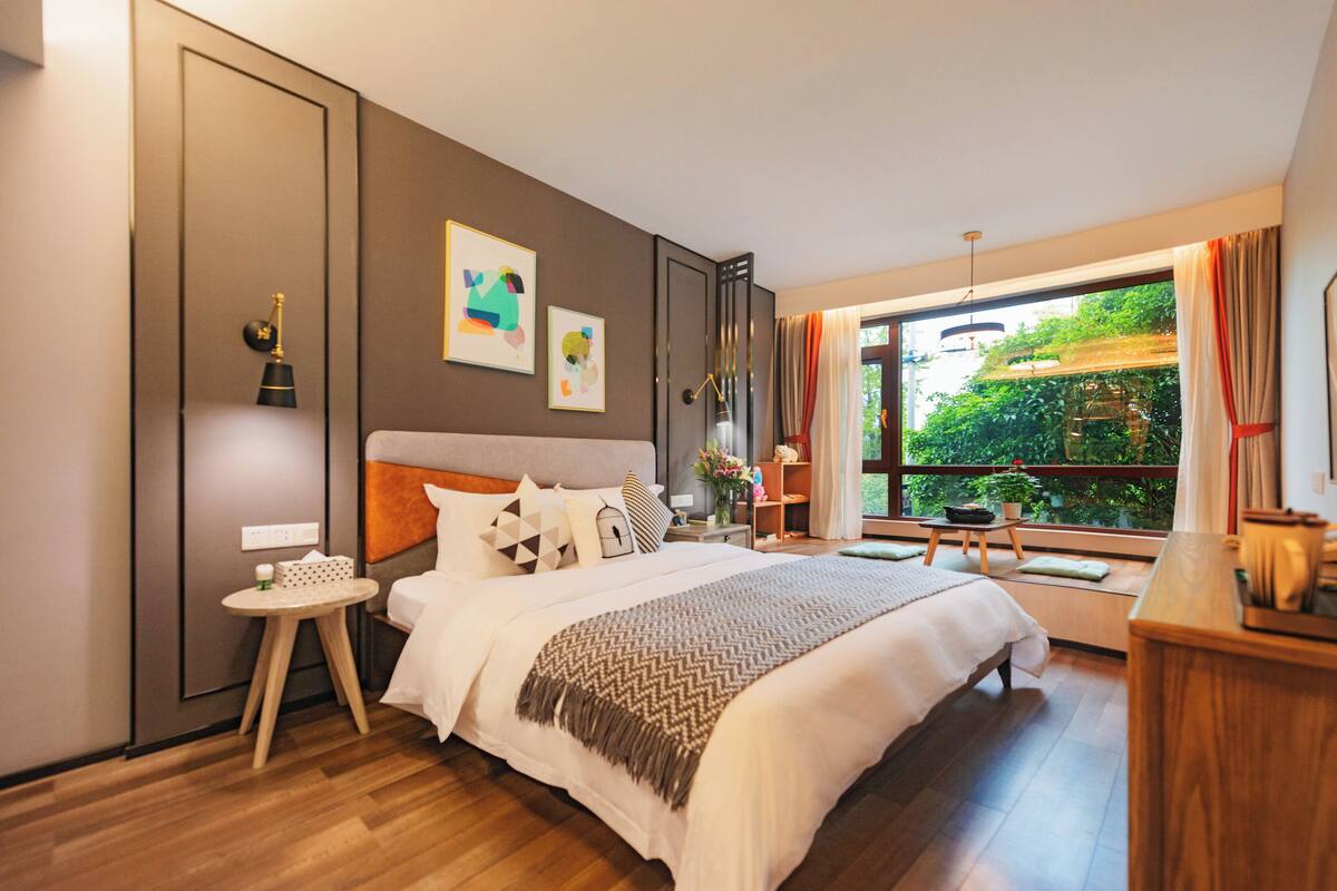 Una habitación de hotel moderna con una cama grande, una mesita auxiliar pequeña, un escritorio con una silla y un ventanal que ofrece vistas a zonas verdes.