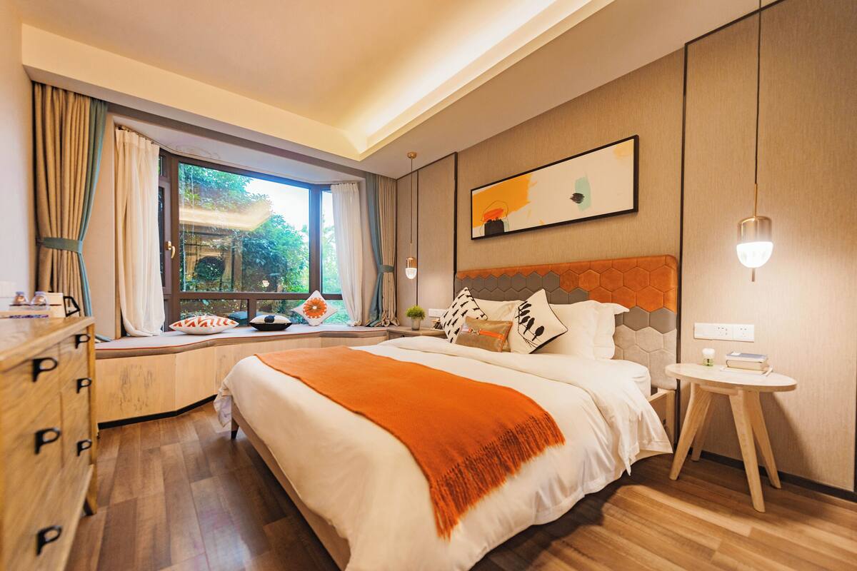 Una habitación de hotel moderna con una cama grande, una ventana con cortinas, una mesita de noche y una obra de arte colgada en la pared.