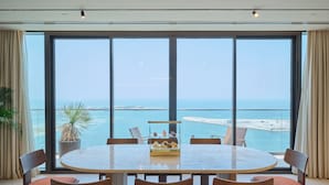 A dining area with a round table and six chairs, large windows with a view of the sea and a beach, and a potted plant on the left. - The OQ (Lusail)