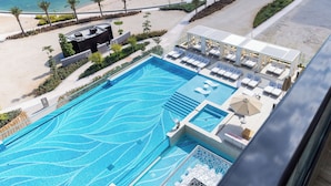 An aerial view of a resort with a large swimming pool, sun loungers, and a beach area. - The OQ (Lusail)
