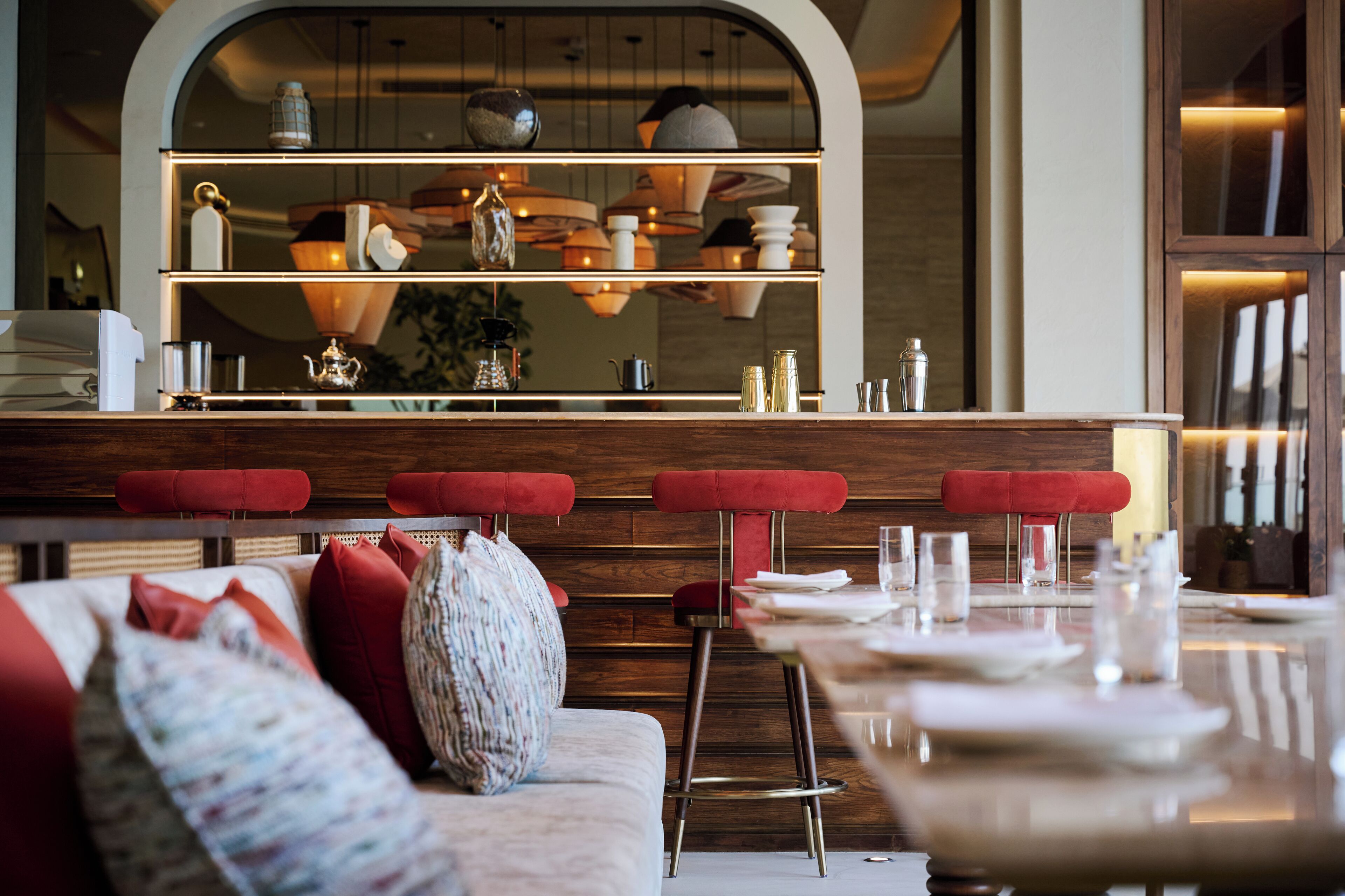 A modern restaurant interior with a bar area, red upholstered stools, and a variety of table settings.