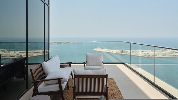 A balcony with two white cushioned chairs and a wooden bench, overlooking a coastal view with a clear sky. - The OQ (Lusail)