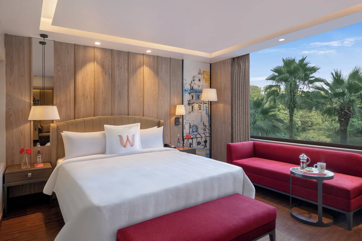 A hotel room with a large bed, a red sofa, a small table, and a view of palm trees.